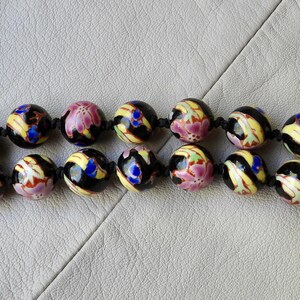 May include: Close-up of a beaded necklace featuring large, round beads. The beads are primarily black with colorful floral designs in pink, yellow, blue, and green. The beads are strung on a black cord.