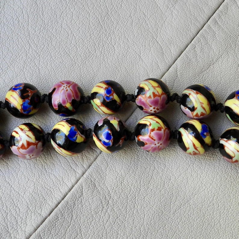 May include: Close-up of a beaded necklace featuring large, round beads. The beads are primarily black with colorful floral designs in pink, yellow, blue, and green. The beads are strung on a black cord.