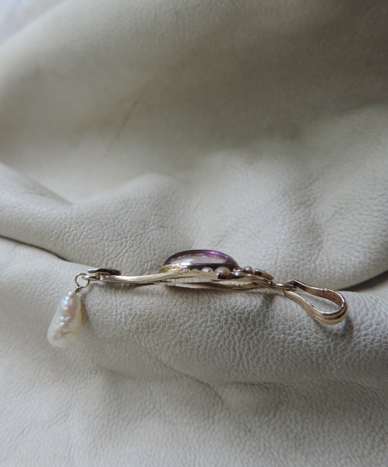 Puede incluir: Colgante dorado con una gran gema ovalada p&uacute;rpura en el centro. El colgante tiene un dise&ntilde;o delicado con peque&ntilde;as gemas blancas redondas y una perla colgante de forma irregular. El cierre es un lazo simple.