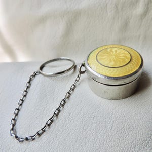 Puede incluir: Pequeña caja redonda de plata con tapa con bisagras. La tapa está decorada con un diseño de esmalte amarillo. Una cadena y un anillo de plata están unidos a la caja. Probablemente un artículo vintage o antiguo.