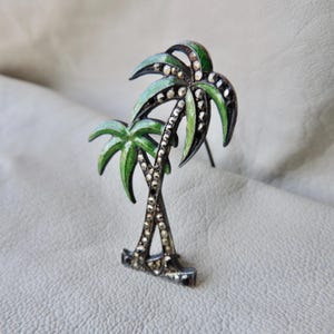 May include: A vintage palm tree brooch with green enamel leaves and sparkling accents. The brooch features two palm trees with detailed fronds and a silver-tone metal base. A tropical-themed accessory.