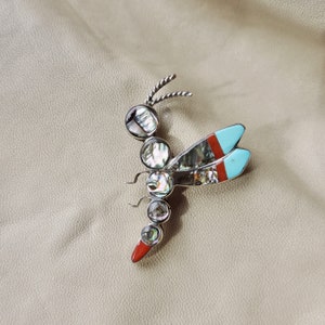 Pode incluir: Broche de libélula prateada com segmentos de corpo de concha de abalone iridescente e detalhes turquesa e vermelhos nas asas. O broche tem um design detalhado e texturizado.