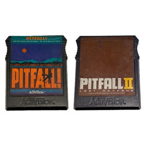 May include: Two black plastic video game cartridges for the Atari 2600. The left cartridge features a blue and orange label with the text PITFALL! and Activision. The right cartridge has a brown label with the text PITFALL II LOST CAVERNS and Activision.