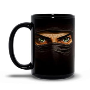 Puede incluir: Taza de cerámica negra con una ilustración detallada de los ojos de una persona mirando a través de una tela negra. Los ojos son verdes y la taza tiene un asa negra.