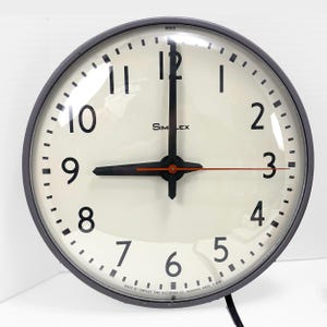 Può includere: Un orologio Simplex rotondo con cornice nera e quadrante color crema. Presenta numeri neri, lancette nere e una lancetta dei secondi rossa. Il quadrante indica "Simplex" e "MADE BY SIMPLEX TIME RECORDER CO. GARDNER, MASS. U.S.A."