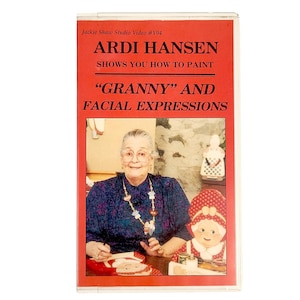 May include: A retro VHS video cassette with a red cover. The text reads "ARDI HANSEN SHOWS YOU HOW TO PAINT 'GRANNY' AND FACIAL EXPRESSIONS." The image shows an elderly woman wearing glasses and a necklace, alongside a painted 'granny' image.