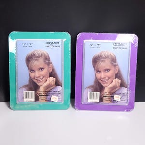 May include: Two plastic picture frames, one green and one purple, both with a 5x7 inch opening. The frames are packaged and ready to be used. The frames are labeled "CRYSTALITE PHOTOFRAME" and have instructions for use.