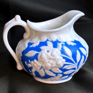 May include: A white and blue creamer jug with a floral pattern. The jug has a white handle and spout, with a blue background and white floral relief. It is likely made of ceramic or porcelain.