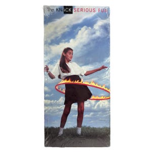Puede incluir: Una cinta VHS de "The Knack: Serious Fun" muestra a una persona joven con camisa blanca y falda negra, haciendo hula hoop con un aro en llamas. El fondo muestra un cielo azul con nubes y hierba verde.