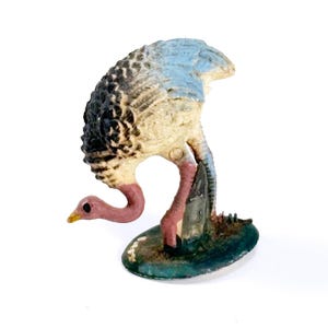 May include: A small, vintage ostrich figurine. The bird has a cream-colored body, a black and brown patterned back, and a light blue head. Its neck and legs are pink, and it stands on a green base.