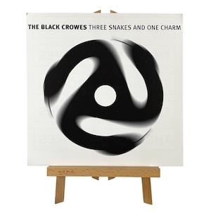 May include: A square album cover for "The Black Crowes: Three Snakes and One Charm" is displayed on a small wooden easel. The cover features a black and white abstract design with a central white circle. The album title is printed in black.