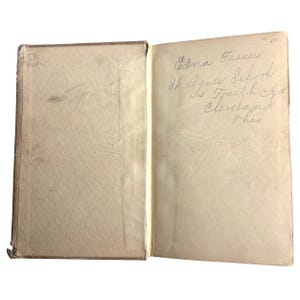 May include: An open vintage book with aged, off-white pages. The left page is blank, while the right page features handwritten text: "Edna Traseu, St. Agnes School, 1115 Fourth Ave, Cleveland, Ohio."