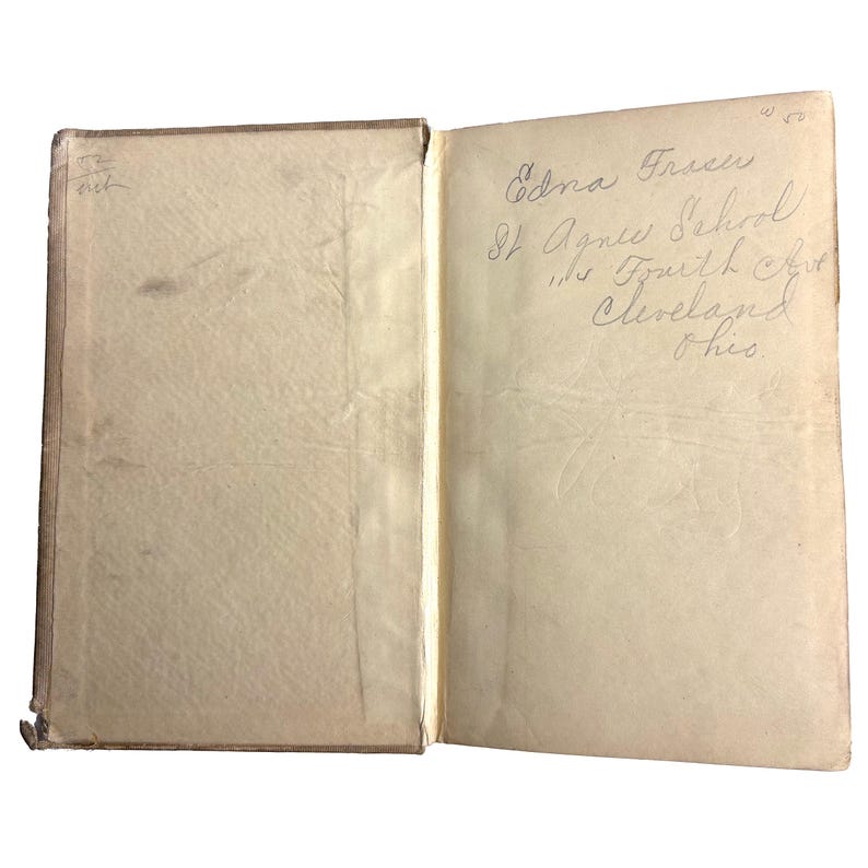 May include: An open vintage book with aged, off-white pages. The left page is blank, while the right page features handwritten text: "Edna Traseu, St. Agnes School, 1115 Fourth Ave, Cleveland, Ohio."
