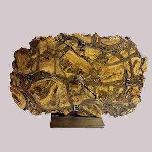 Peut inclure: Une horloge de bureau unique, fabriquée à partir d'une grande pièce de pierre de forme irrégulière. Le cadran de l'horloge présente un motif naturel et moucheté dans des tons de marron et d'or, avec des chiffres et des aiguilles noirs. L'horloge est montée sur une base métallique rectangulaire.