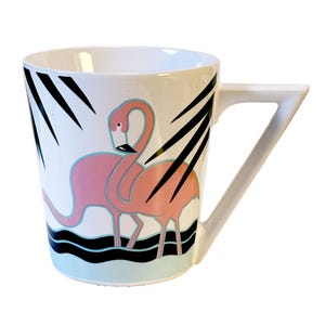 May include: White ceramic mug featuring a pink flamingo design with black accents. The mug has a unique handle and is decorated with black palm fronds and a light blue wave pattern at the base. A fun and colorful addition to any kitchen.