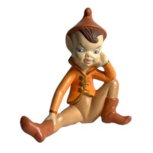 Könnte beinhalten: Vintage Keramikfigur eines sitzenden Elfen. Der Elf hat einen hellen Hautton und trägt eine orangefarbene Jacke mit goldenen Knöpfen, braune Stiefel und einen passenden Hut. Der Elf stützt seinen Kopf auf seine Hand.