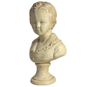 May include: A cream-colored bust of a young person with short, wavy hair and a serene expression. The sculpture features a detailed collar and is mounted on a pedestal base. The bust is made of a light-colored material.