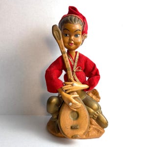 Puede incluir: Una figura vintage de un músico con un atuendo y sombrero rojos, sosteniendo un laúd. La figura tiene una cara pintada y detalles dorados. La base es de color marrón claro.