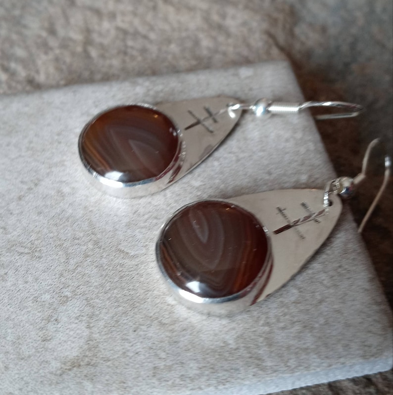 May include: A pair of silver dangle earrings with brown agate stones. The stones have a swirling pattern and are set in a silver teardrop shape. The earrings have a simple design and are perfect for everyday wear.