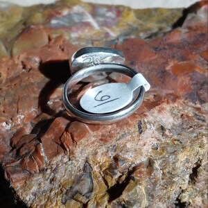 K&ouml;nnte beinhalten: Ein silberner Ring mit einem runden, grauen Stein, der in die Band gefasst ist. Der Ring liegt auf einem St&uuml;ck rotem und braunem Gestein. Der Ring ist mit der Nummer "6" gekennzeichnet.