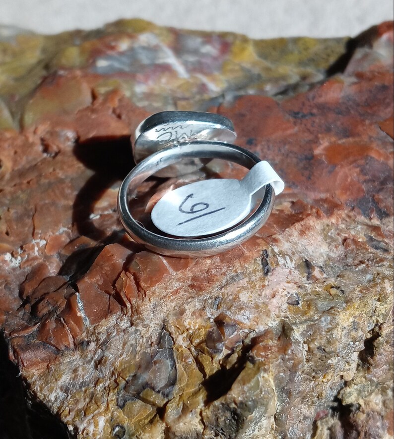 K&ouml;nnte beinhalten: Ein silberner Ring mit einem runden, grauen Stein, der in die Band gefasst ist. Der Ring liegt auf einem St&uuml;ck rotem und braunem Gestein. Der Ring ist mit der Nummer "6" gekennzeichnet.