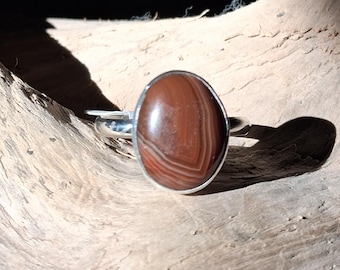 May include: A silver ring featuring a smooth, oval-shaped brown agate gemstone with distinct concentric banding patterns. The stone is set in a polished silver bezel, and the band is a simple, rounded silver wire that encircles the finger.
