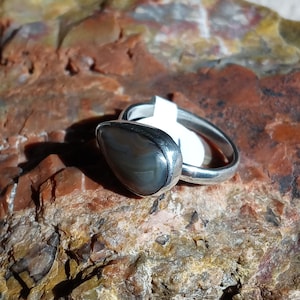 K&ouml;nnte beinhalten: Ein silberner Ring mit einem grauen und braunen Achat-Stein, der in einer Zargenfassung sitzt. Der Ring liegt auf einem felsigen Untergrund.