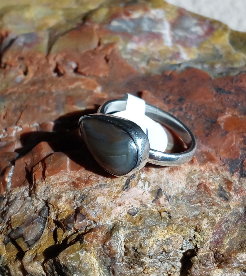 K&ouml;nnte beinhalten: Ein silberner Ring mit einem grauen und braunen Achat-Stein, der in einer Zargenfassung sitzt. Der Ring liegt auf einem felsigen Untergrund.