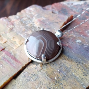 May include: A silver pendant with a brown and white agate stone. The stone has a swirling pattern and is set in a silver bezel.