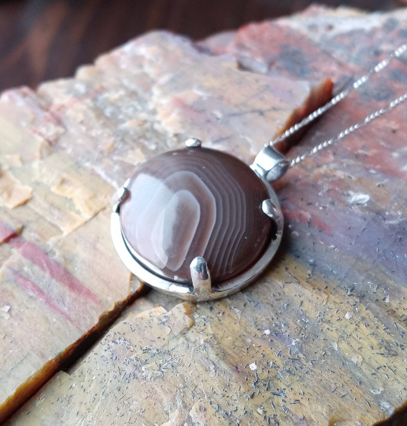 May include: A silver pendant with a brown and white agate stone. The stone has a swirling pattern and is set in a silver bezel.