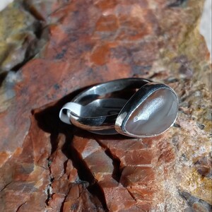 K&ouml;nnte beinhalten: Ein silberner Ring mit einem grauen Achat-Stein, der in einer Zargenfassung gefasst ist. Der Ring liegt auf einem braunen und grauen Gesteinsuntergrund.