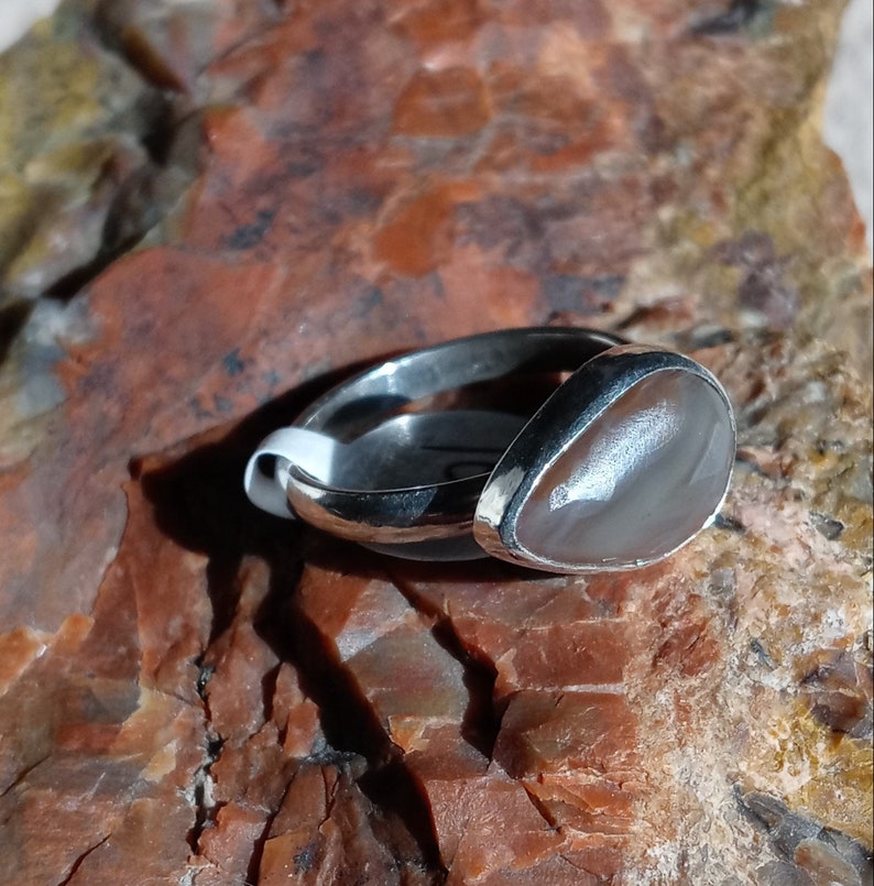K&ouml;nnte beinhalten: Ein silberner Ring mit einem grauen Achat-Stein, der in einer Zargenfassung gefasst ist. Der Ring liegt auf einem braunen und grauen Gesteinsuntergrund.