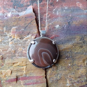 May include: A silver chain necklace with a round pendant featuring a brown and white agate stone set in a silver bezel.