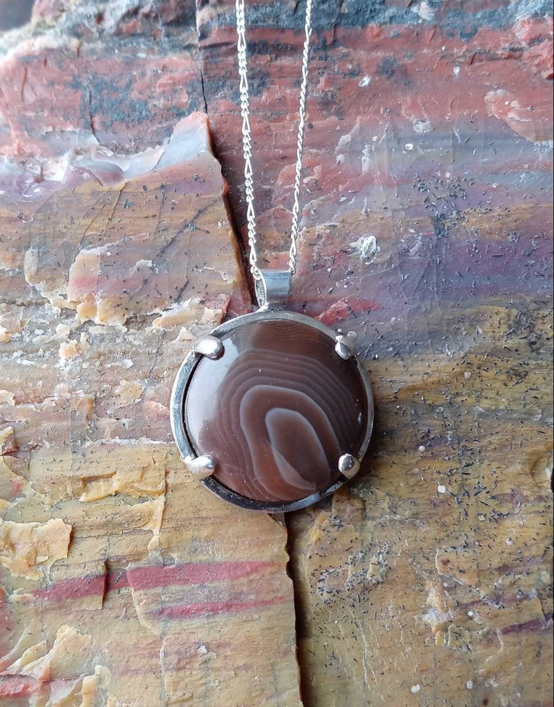 May include: A silver chain necklace with a round pendant featuring a brown and white agate stone set in a silver bezel.
