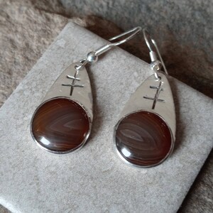 May include: A pair of silver dangle earrings with brown agate stones. Each earring features a silver teardrop shape with a cross design.