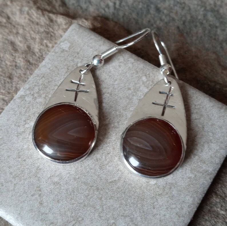 May include: A pair of silver dangle earrings with brown agate stones. Each earring features a silver teardrop shape with a cross design.