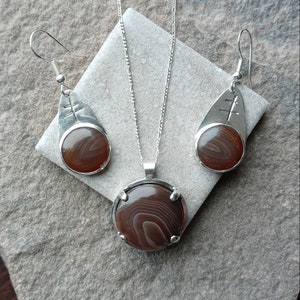 May include: A silver necklace and earrings set with brown agate stones. The necklace features a round pendant with a silver frame and the earrings are teardrop shaped with a silver frame.