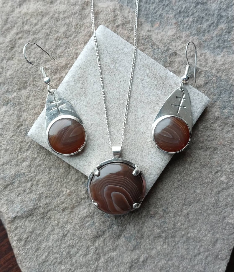 May include: A silver necklace and earrings set with brown agate stones. The necklace features a round pendant with a silver frame and the earrings are teardrop shaped with a silver frame.