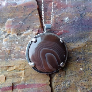 May include: A silver pendant necklace with a brown agate stone set in a silver bezel. The stone has a swirling pattern of brown and white.