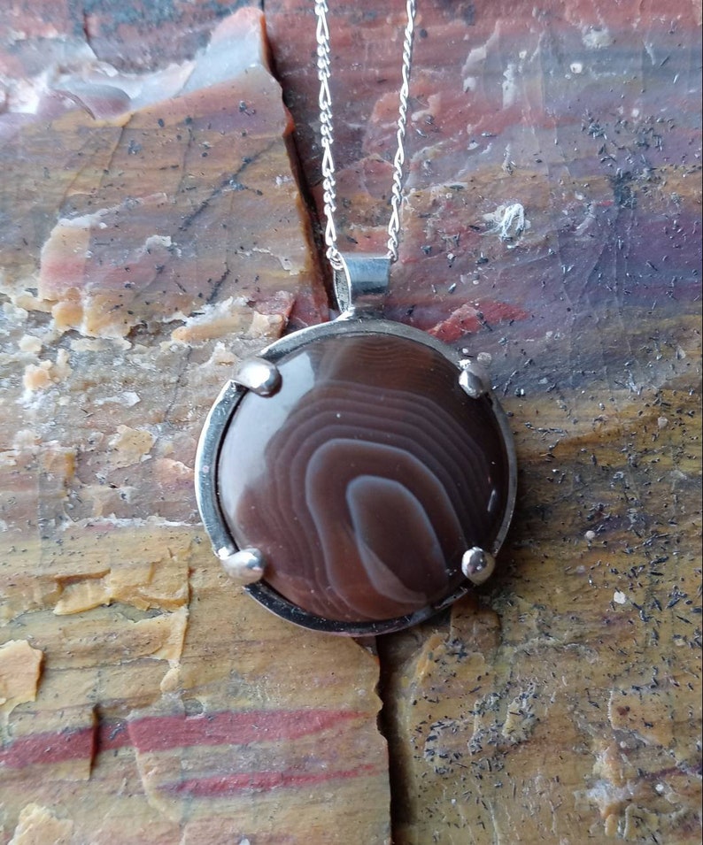 May include: A silver pendant necklace with a brown agate stone set in a silver bezel. The stone has a swirling pattern of brown and white.
