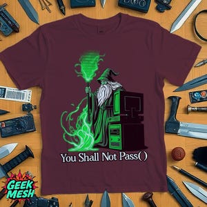 Op de afbeelding: Bordeaux t-shirt met een afbeelding van een tovenaar met een lange witte baard en een groene mantel, die een staf vasthoudt die groene binaire code uitstraalt. Een computer staat naast de tovenaar. De tekst "You Shall Not Pass()" staat eronder. Het merklogo "Geek Mesh" staat onderaan.