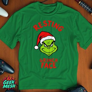 May include: Green t-shirt featuring a Grinch face wearing a Santa hat. The text "RESTING" is above the Grinch's face, and "GRINCH FACE" is below it, all in red. The shirt is a short-sleeved crew neck.