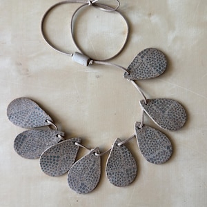 May include: A necklace featuring teardrop-shaped pendants with a textured, speckled pattern in shades of brown and gray. The pendants are strung on a thin, light brown cord with an adjustable closure. Jewelry.