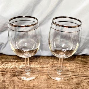 Peut inclure: Deux verres à vin transparents avec un détail de bordure doré. Les verres sont posés sur une surface en bois avec un tissu bleu clair en arrière-plan. La verrerie est élégante et adaptée au service du vin.
