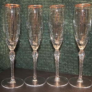 May include: Four clear champagne flutes with gold rims and decorative stems. The flutes are arranged in a row, showcasing their elegant design. The stems have a twisted design, adding to their visual appeal. The glasses are on a dark surface.