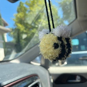 May include: A yellow and black fuzzy bee car charm with white pom-poms for antennae hanging from the rearview mirror of a car.