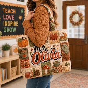 May include: A quilted tote bag featuring a patchwork design with autumn-themed embroidered motifs including pumpkins, mushrooms, a slice of pie, a candle, and a stack of books. The center displays the name Ms. Olivia in bold orange lettering.