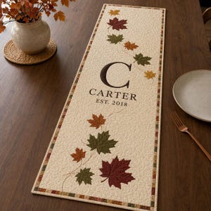 Peut inclure: Un chemin de table matelassé rectangulaire sur fond crème avec une bordure de petits carrés multicolores. Le chemin de table présente des feuilles d'érable d'automne brodées en rouge, orange et vert, avec le texte C CARTER EST. 2018.