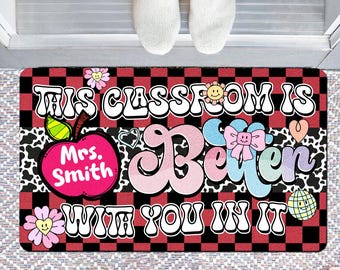 May include: A rectangular doormat with a red and black chequered pattern. The mat features the text "THIS CLASSROOM IS BETTER WITH YOU IN IT" in a retro font, along with an apple graphic and the name "Mrs. Smith". Decorative elements include flowers, hearts, and a smiling sun.