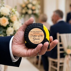 May include: A black and yellow tape measure with a wooden face that reads "No one measures up to you, Father of the Groom, 23.08.23." The tape measure is held in a person's hand, with a wedding ceremony in the background.